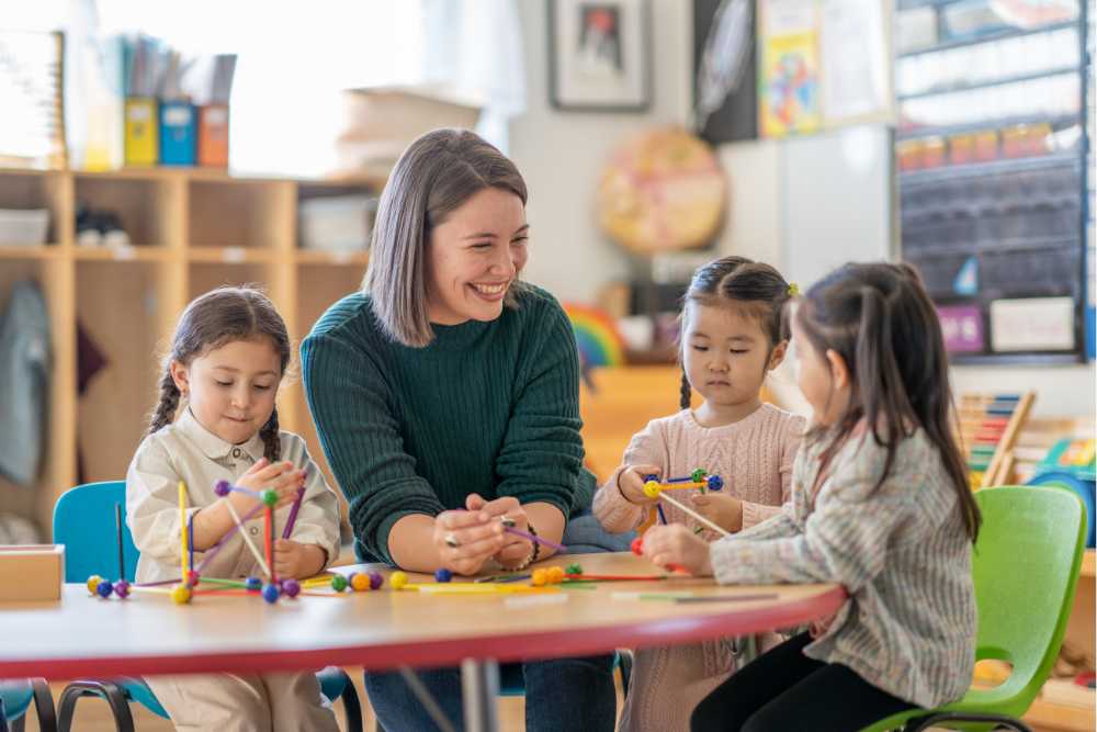 Pedagogisch professional zit aan tafel met spelende kinderen en maakt contact met een kind 